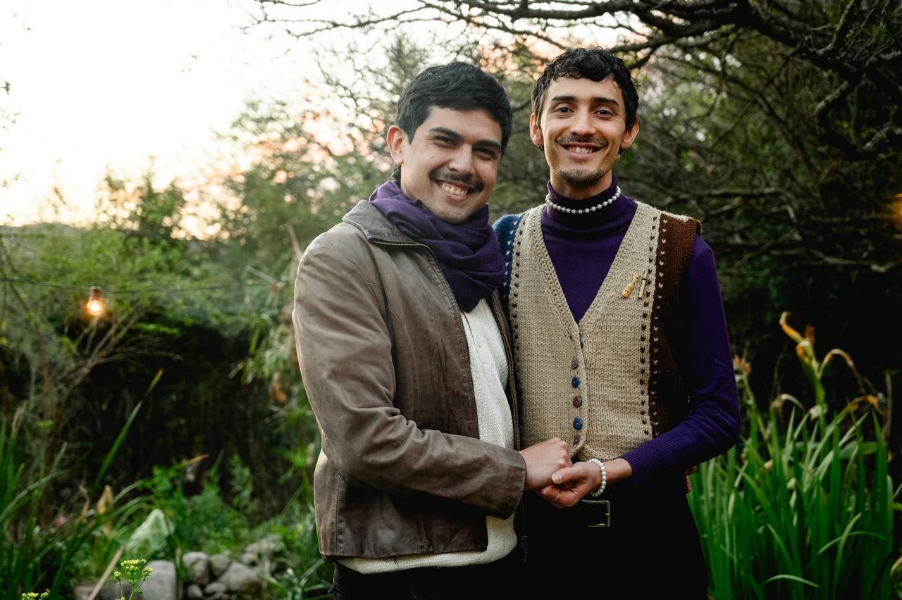 Los dos integrantes de Fulgura Dúo sonríen tomados de la mano en un patio con árboles y plantas.